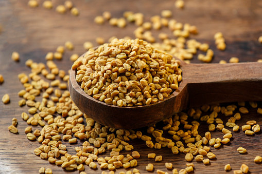 Fenugreek seeds in a wooden spoon and spilling over onto a wooden surface