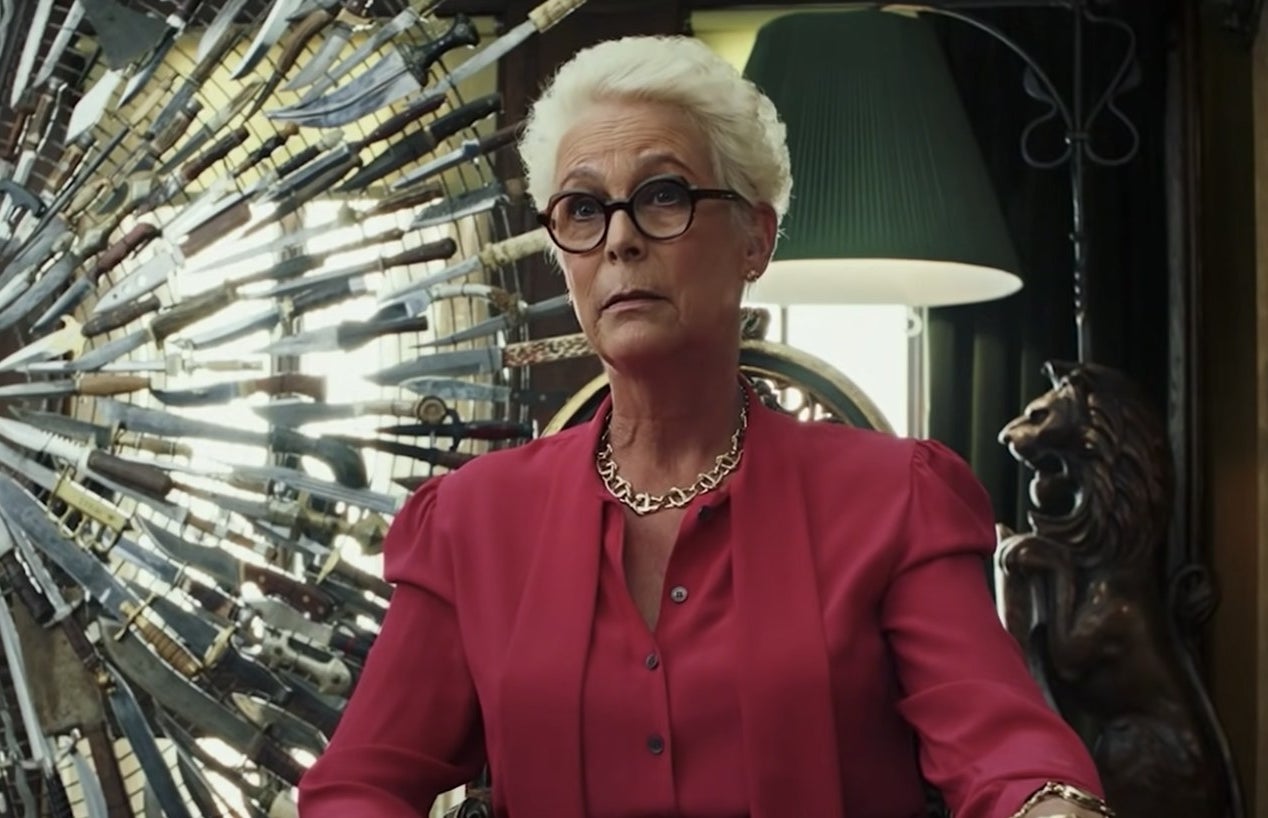 Jamie Lee Curtis in a red suit in front of the decorative wheel of knives from Knives Out.
