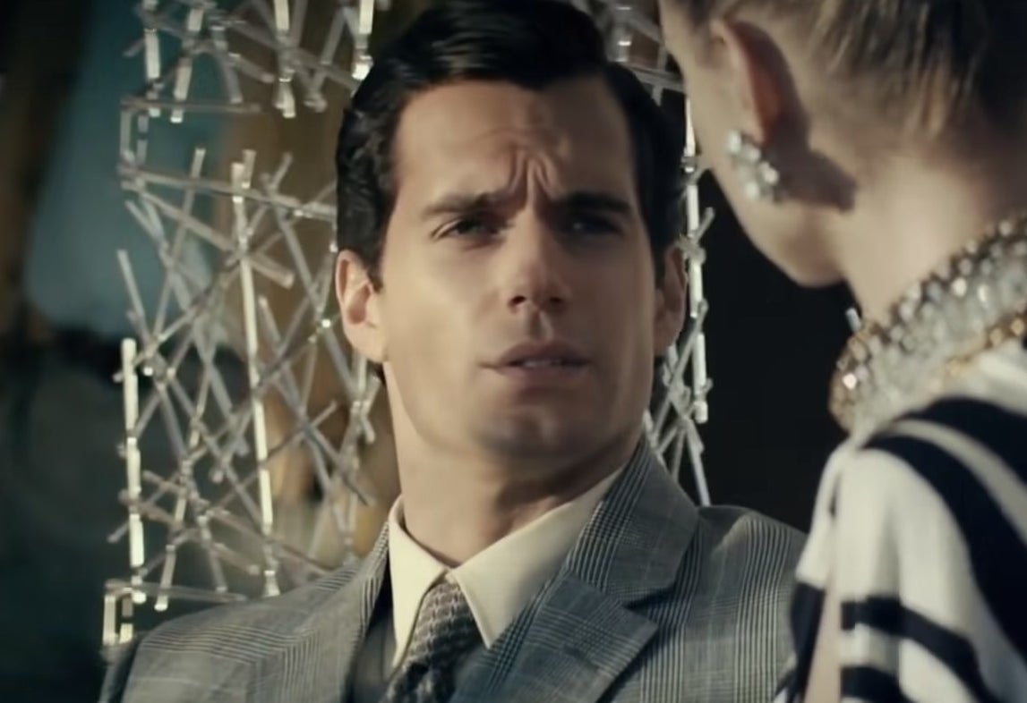 Henry Cavill in a grey, pinstripe suit, talks to a woman who faces away from the camera.