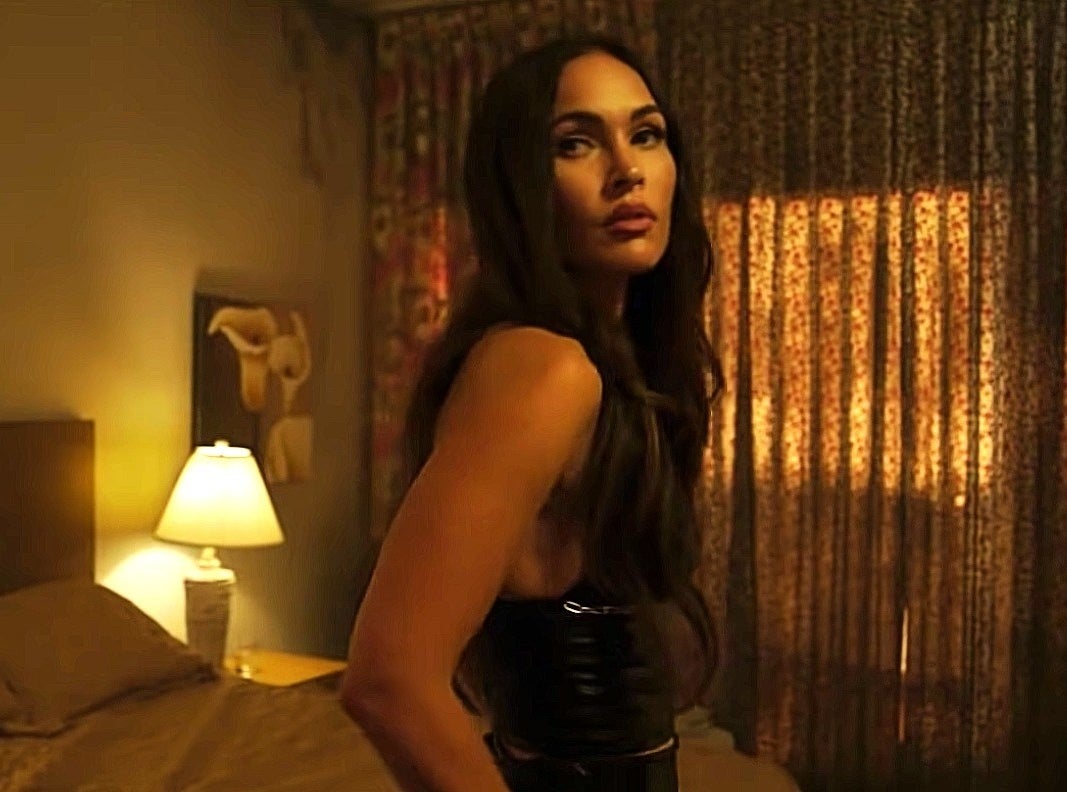 Megan Fox in a black dress stands opposite a bedframe.