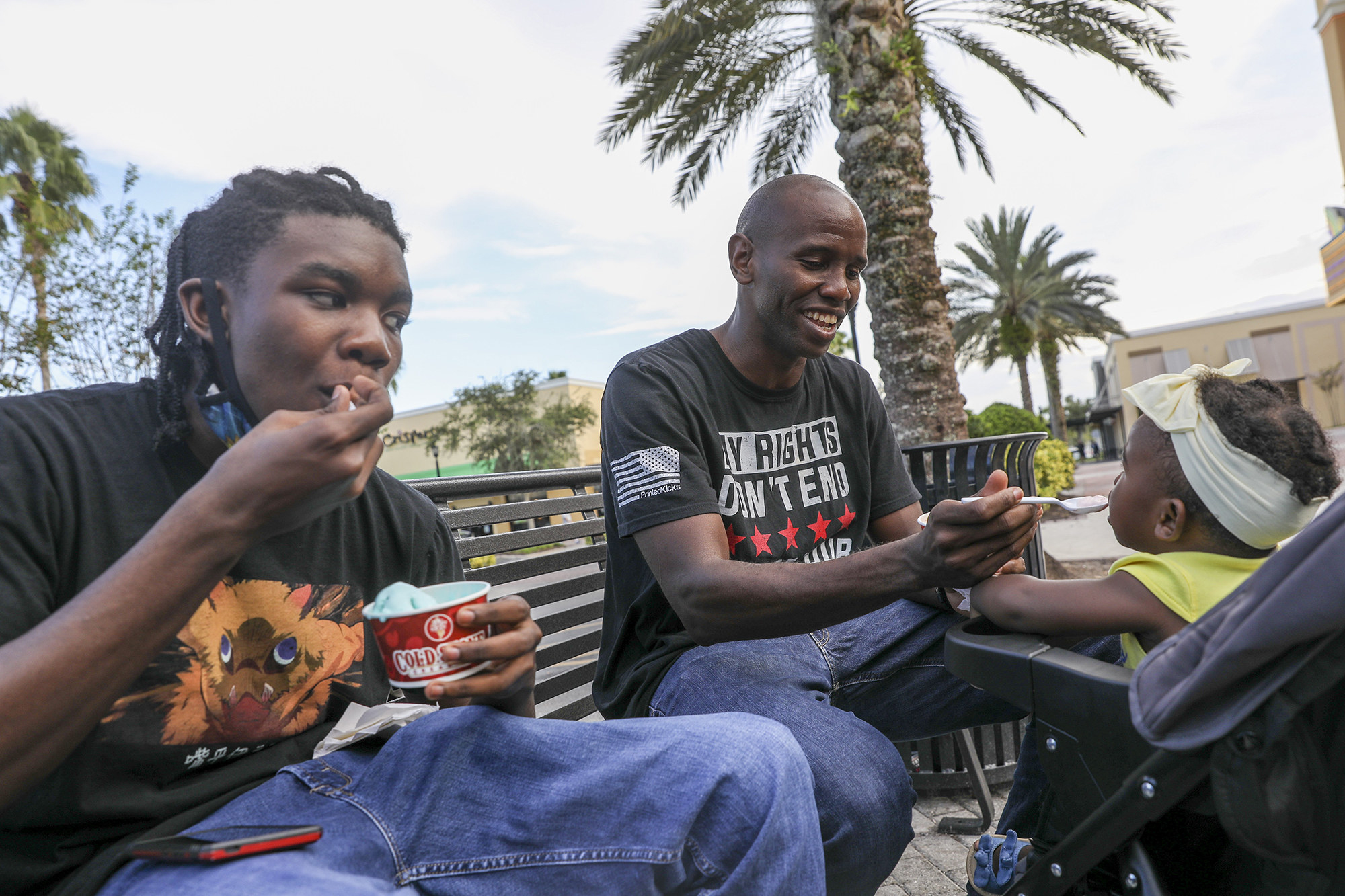 Jerry Wyche spoonfeeds a toddler and sits on a bench next to a teenager eating Cold Stone ice cream and his two children