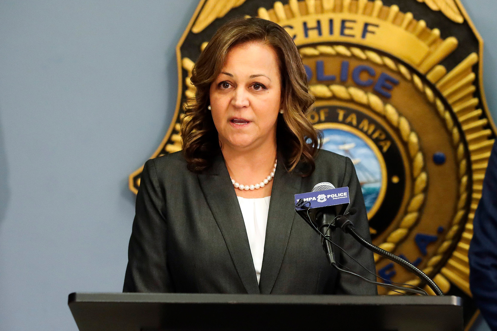 Tampa Police Chief Mary O&#x27;Connor stands behind a lectern and microphone in front of a large police badge decal on the wall