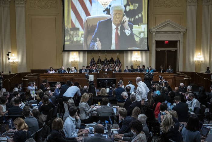 A projected image of Donald Trump appears on a screen above a panel of people in a packed room in the Capitol