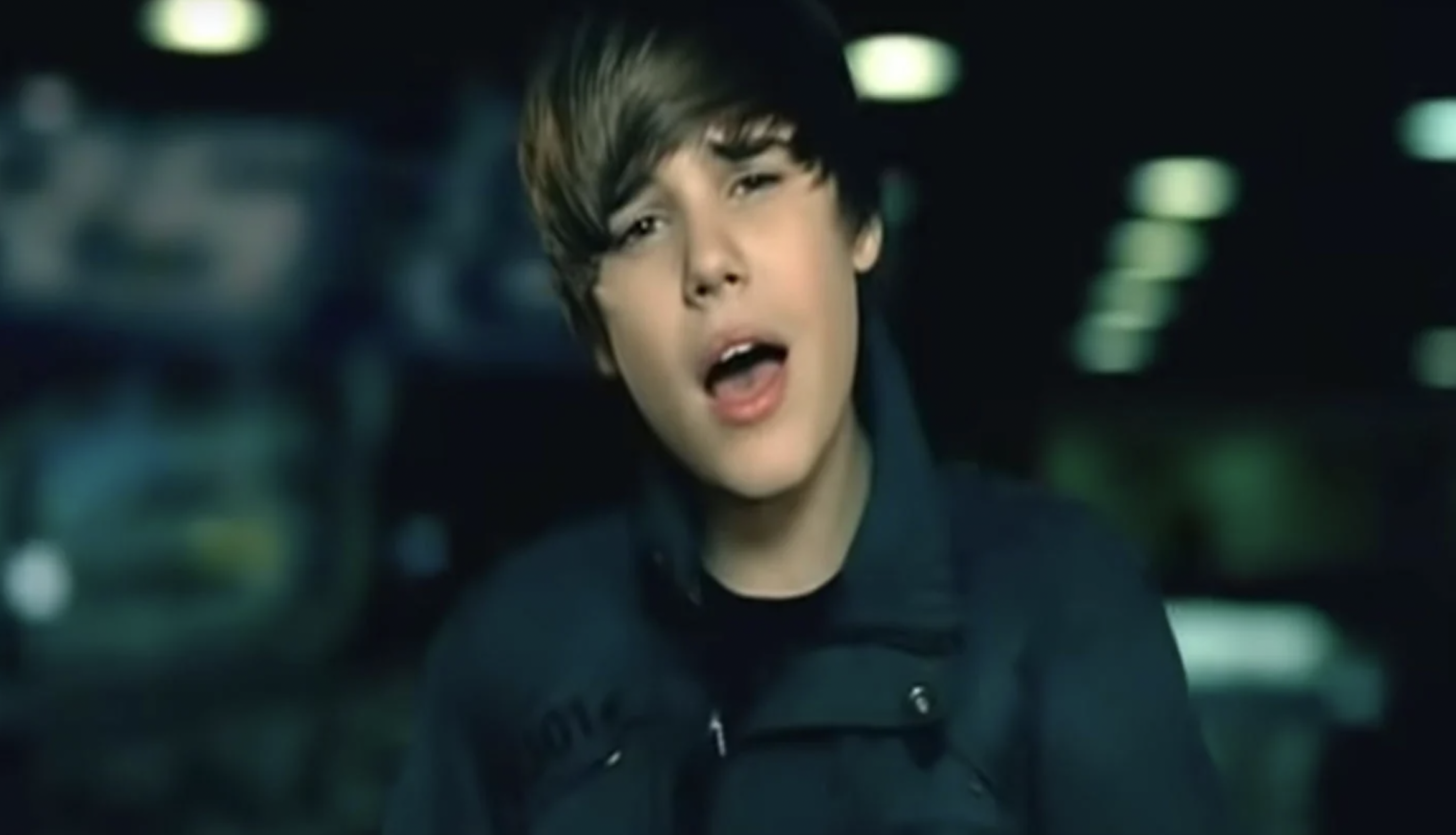 A young Justin Bieber sings while looking at the camera, a blurry night time street is behind him