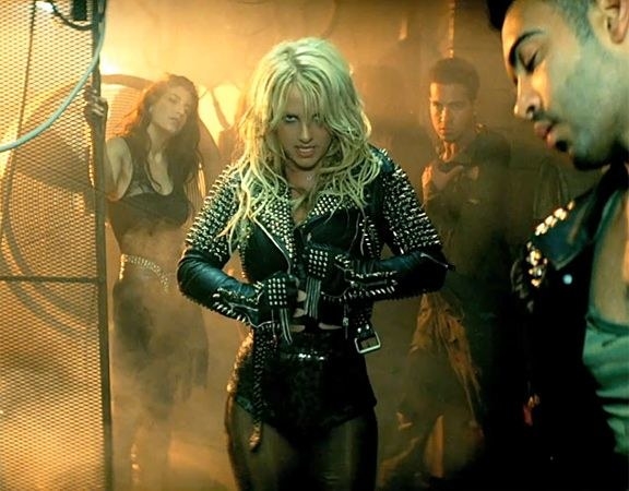 Britney Spears wears a black leather studded jacket on the set of a music video, looking at the camera