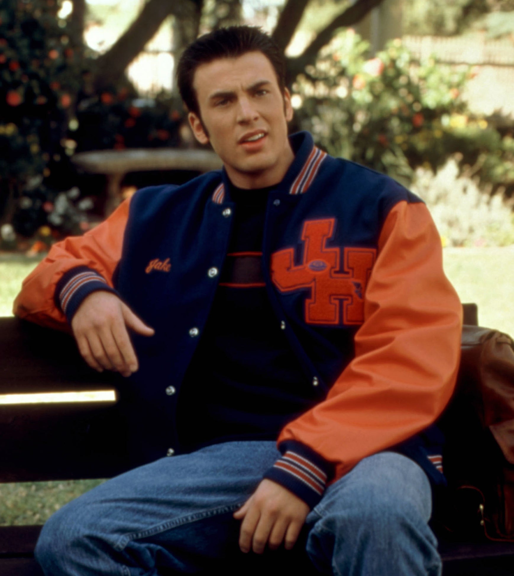 A young Chris Evans wearing a letterman jacket and sitting on a bench