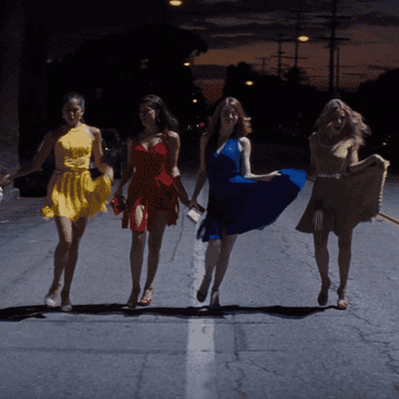 Four women wearing different colored dresses dance down the street