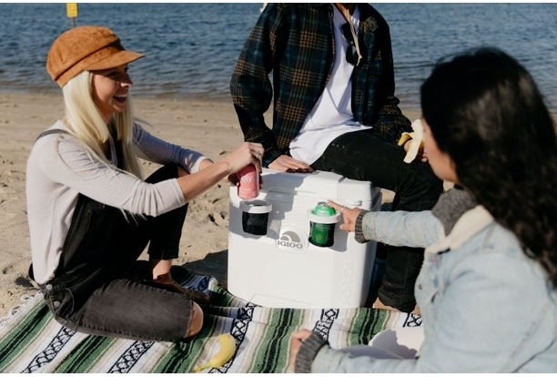 Three people enjoying drinks on a blanket next to white ice chest