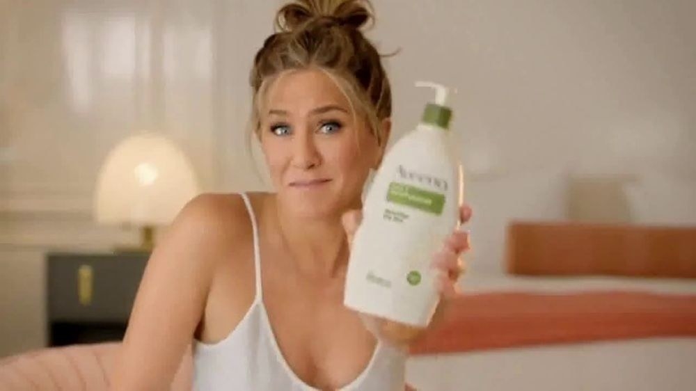 A still from an advertisement of Jennifer Aniston holding up a big bottle of Aveeno moisturizer