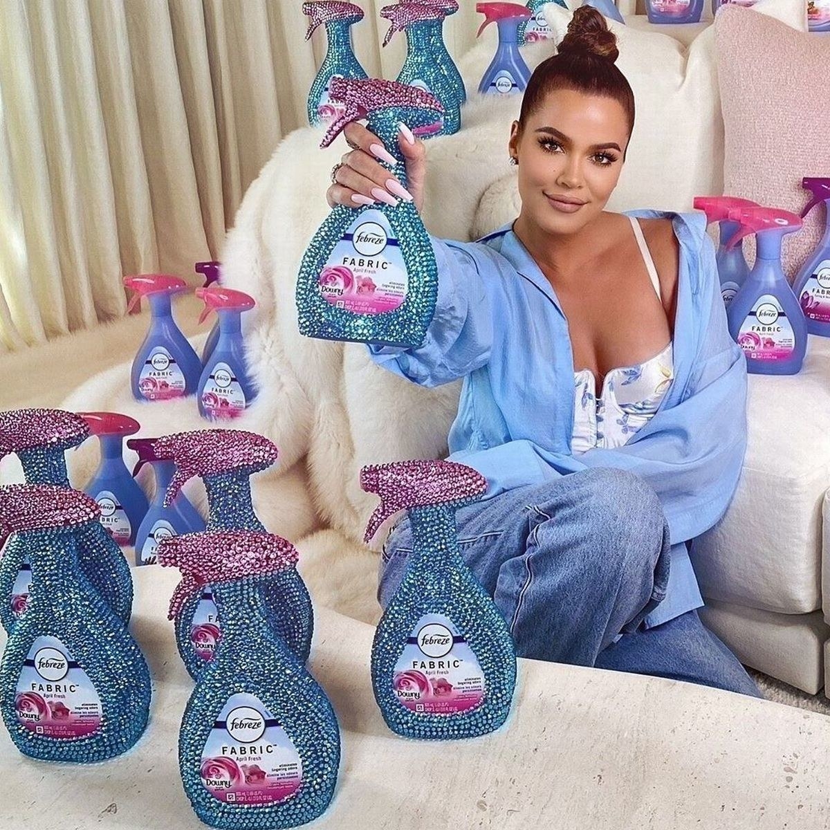 Khloe Kardashian sits in front of a white chair surrounded by spray bottles of Febreze fabric spray, some of which are bejewleled. She holds one out to the camera while smiling