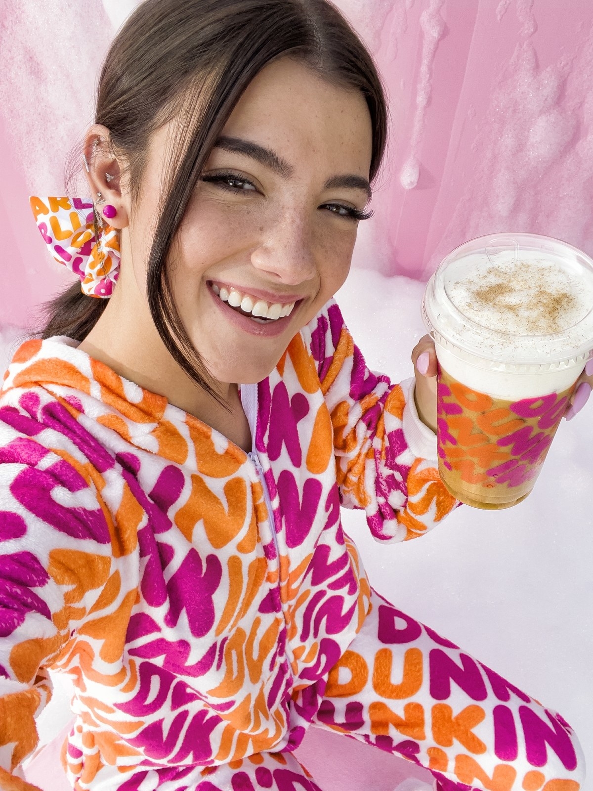 Charli D&#x27;Amelio poses for a selfie in matching Dunkin Donuts pants and jacket, while holding a Dunkin-branded iced coffee