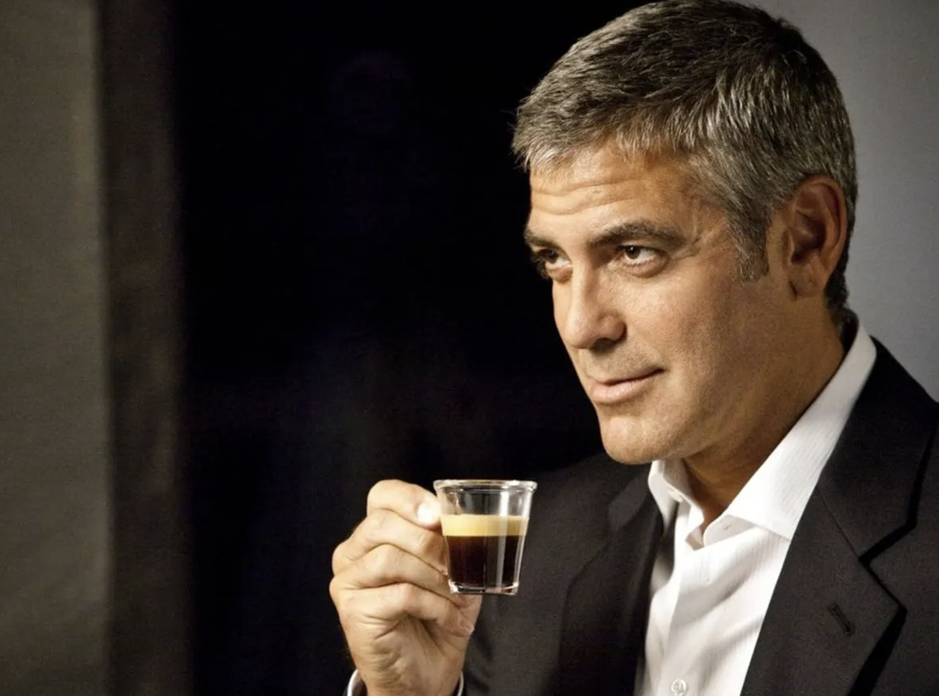 George Clooney holds an espresso shot in a small glass, which he holds up to his face as he looks off mysteriously