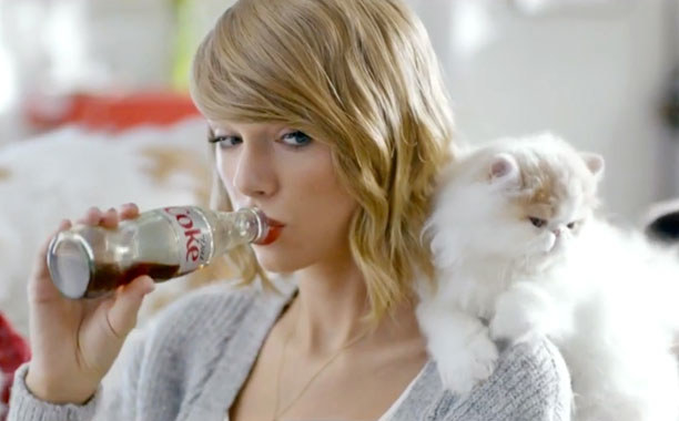 Taylor Swift sips from a glass bottle of Diet Coke while looking at a white fluffy cat on her shoulder