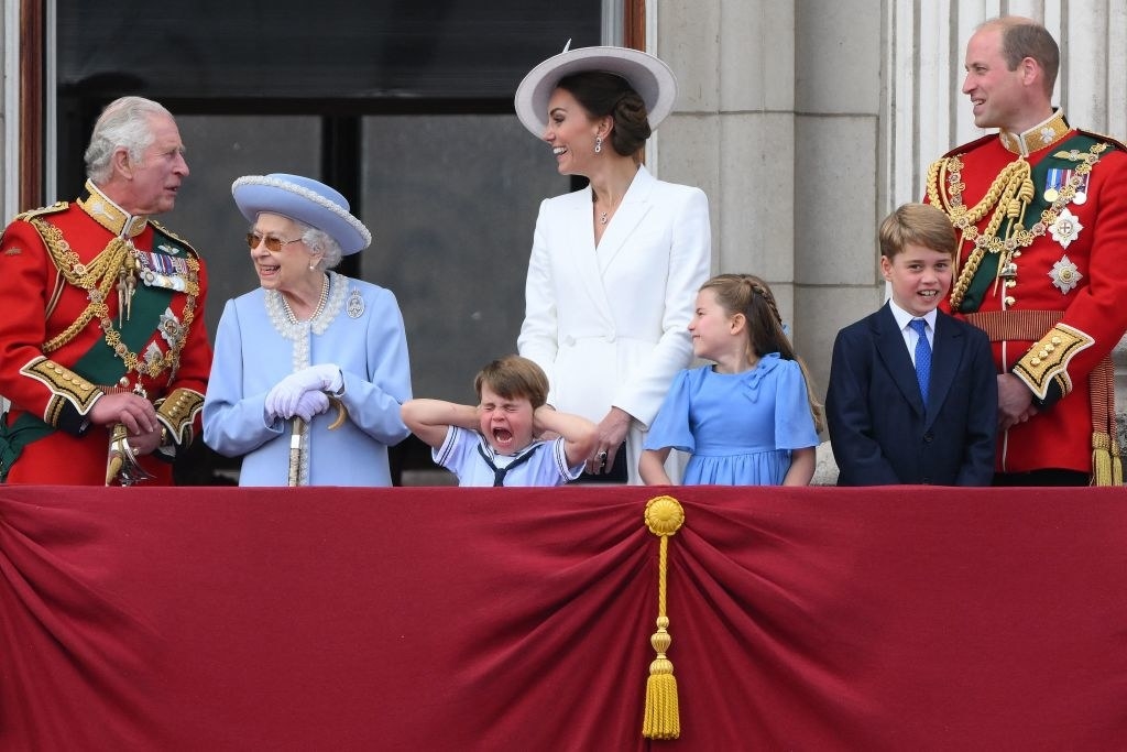 A wide shot of the king and queen, William and Kate, and their other two kids all looking normal, while Louis is screaming with his hands over his ears