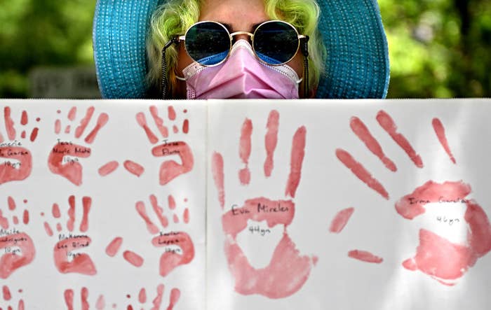 A person wearing sunglasses and a mask stands behind a handmade poster with red hand prints, each written with the name of a victim of the Uvalde school shooting
