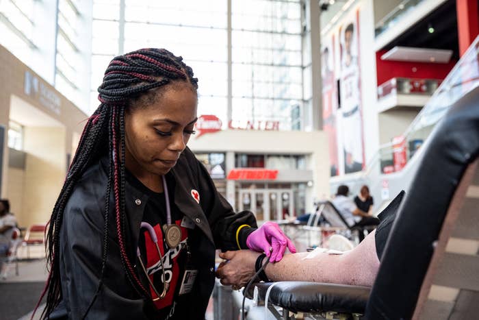 A phlebotomist wearing gloves stands beside someone lying in a recliner with a needle in their arm