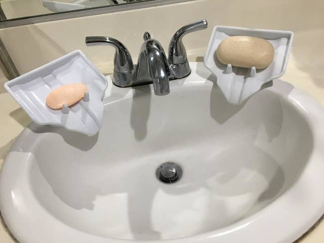 two of the white soap dishes on the side of reviewer's sink. they're  angled downward with little poles to keep the soap in place but also a channel to funnel residue downward