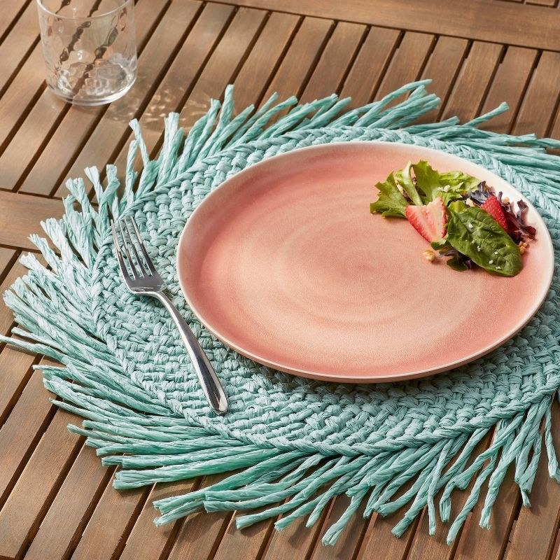 Teal colored fringe placemat under pink plate with small salad on it