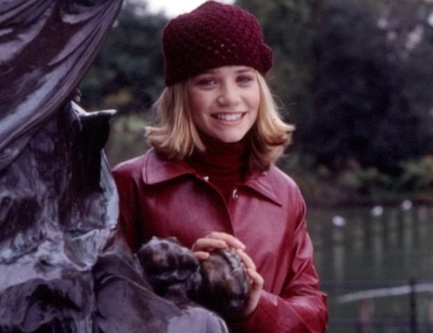 Mary Kate smiles while posing next to a statue wearing a coat and hat