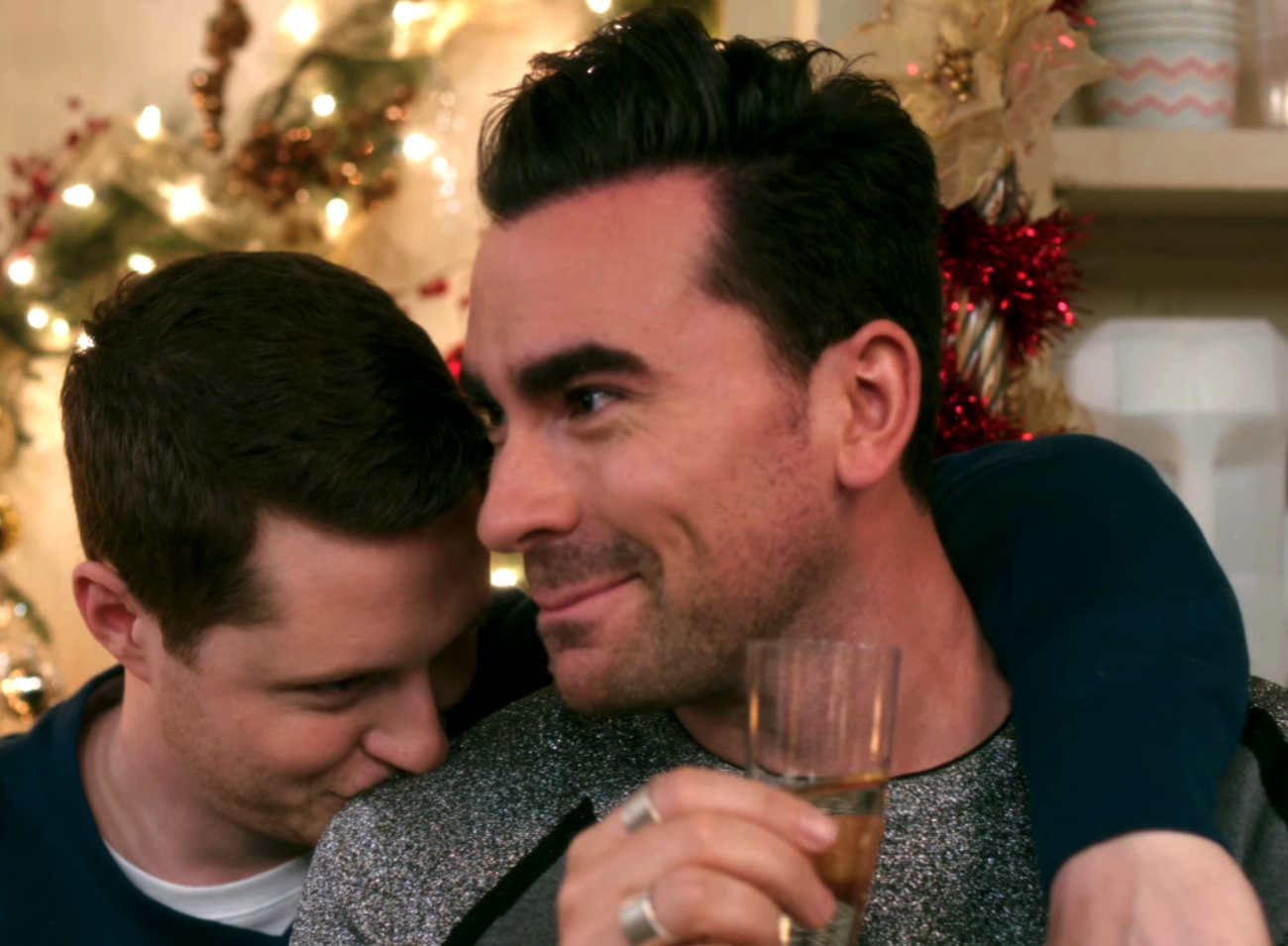 David holding a glass of wine and leaning towards Patrick who is giving him a kiss on his shoulder