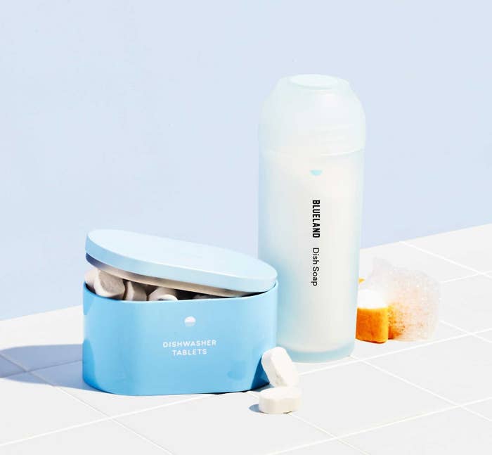 A blue tin of white tablets, a light blue bottle and a yellow sponge with suds on it