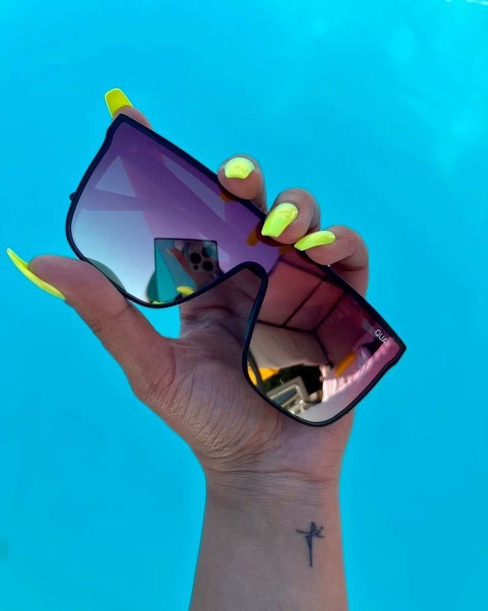 a person&#x27;s hand holding up a pair of the square sunglasses against a bright summery sky