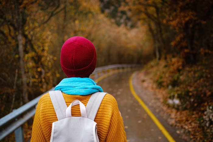 A woman wearing a red beanie and yellow sweater walks along a forest road
