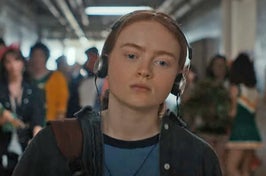 Max from Stranger Things walking down the school hallway with headphones over her ears and a frown on her face