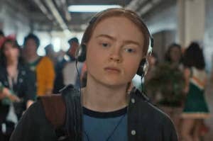 Max from Stranger Things walking down the school hallway with headphones over her ears and a frown on her face