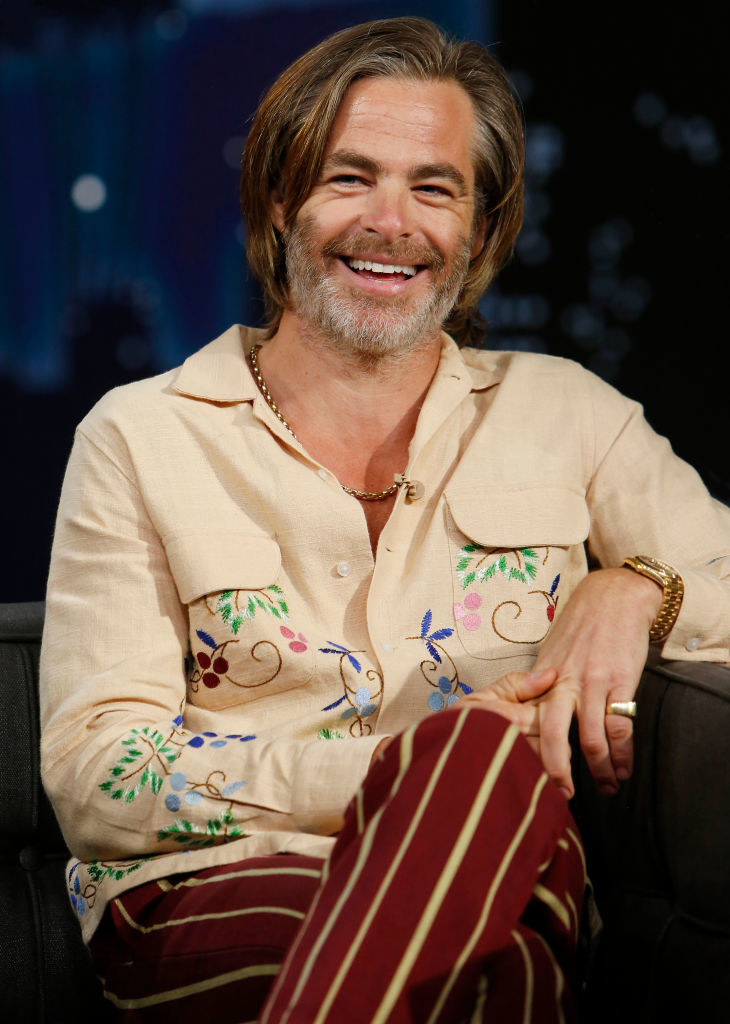 Smiling with longer hair and a beard and mustache and legs crossed