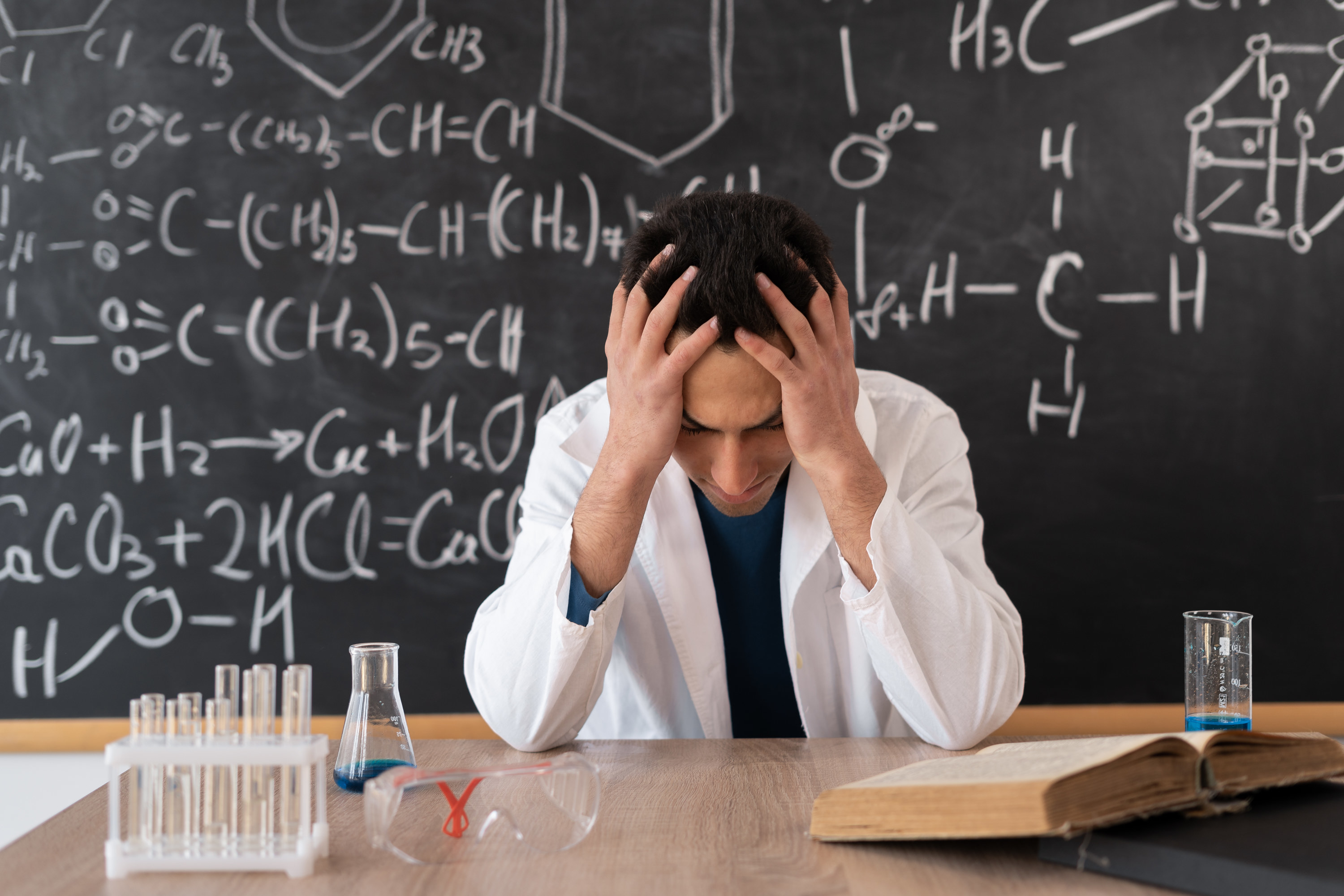 A young Arab teacher sits in a white coat against the background of a blackboard with chemical formulas feeling stressed