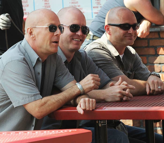 Bruce Willis, wearing sunglasses, sitting next to two other bald men with sunglasses