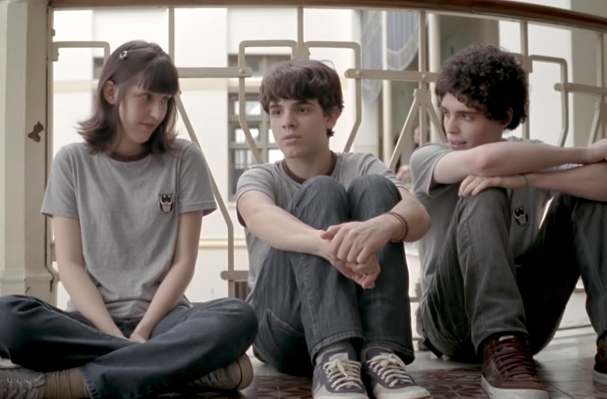Three teens in school uniforms sit on the floor in a row in conversation