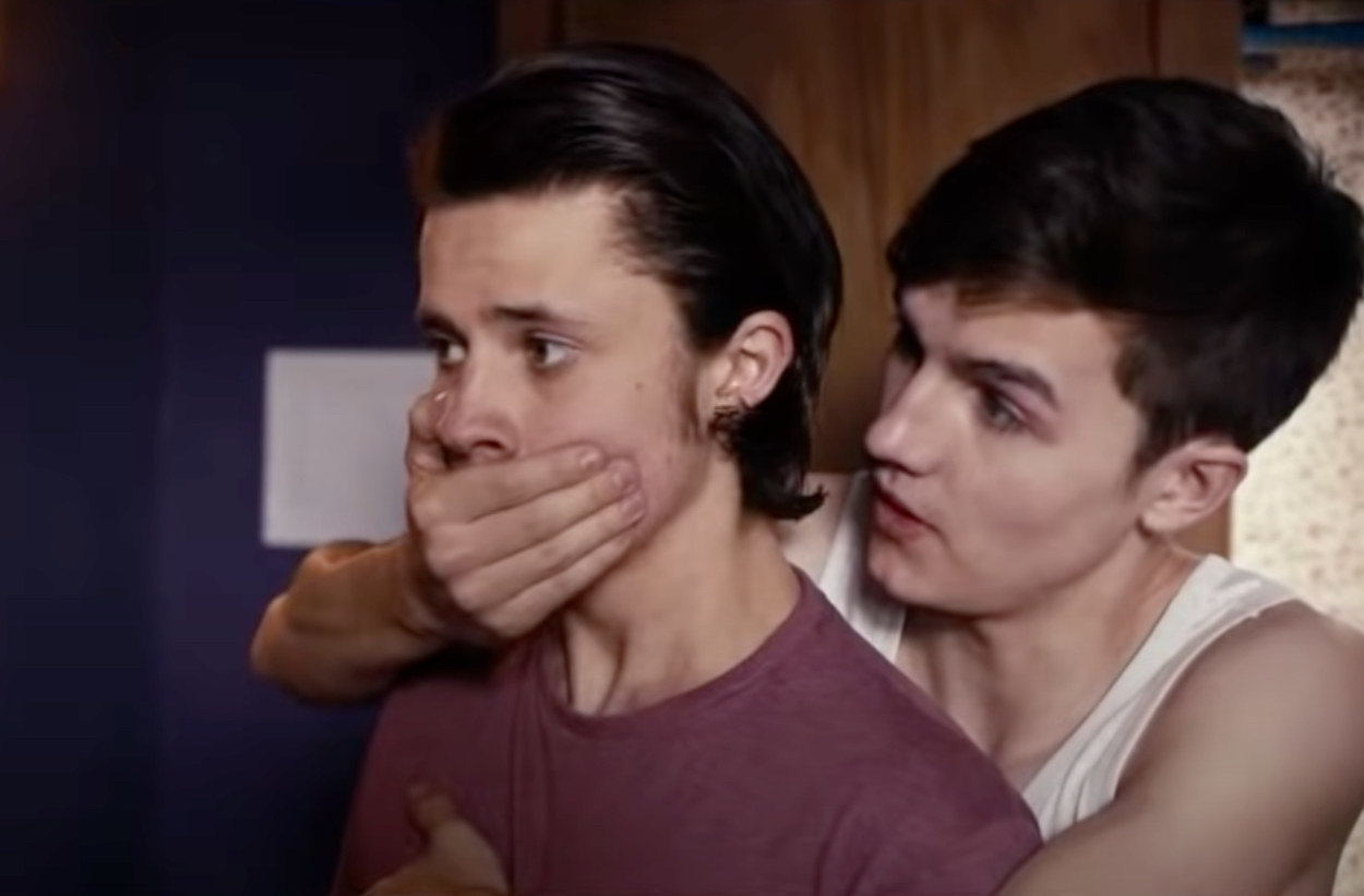 A teenager boy stands behind another teenage boy holding him up against him and covering his mouth in a bedroom