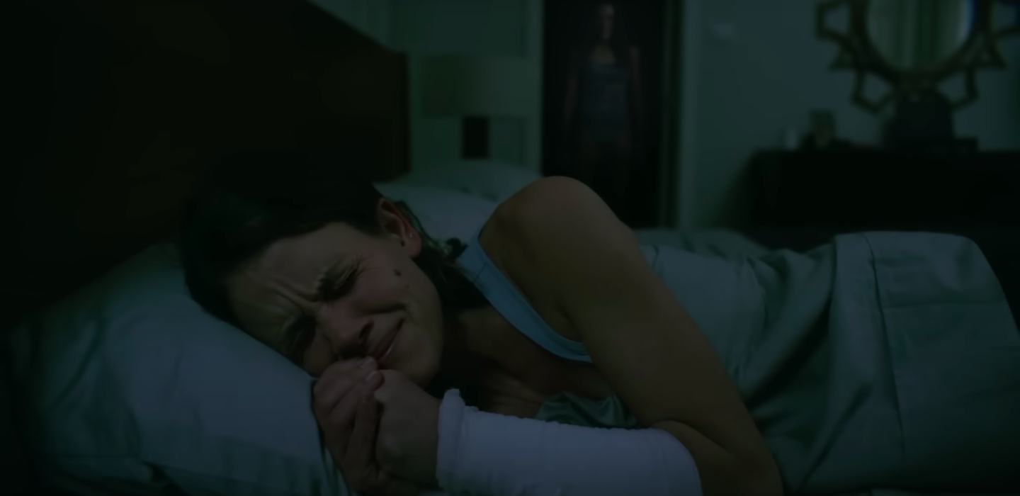 A woman is terrified in bed as a woman approaches from behind in the shadows