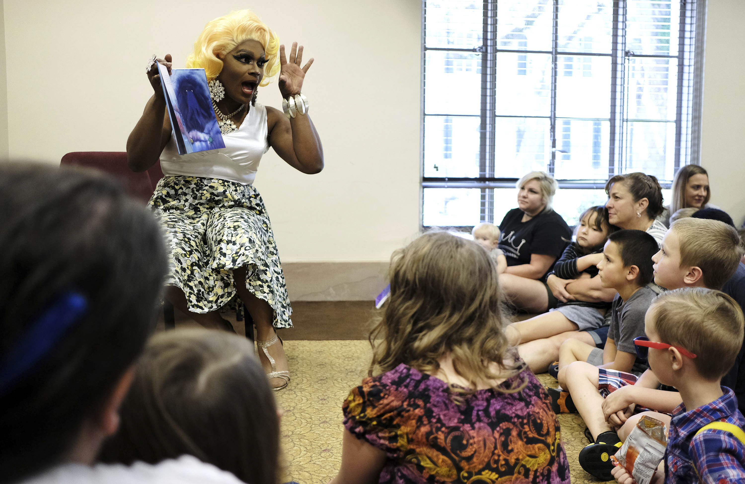 A person holding up a book and wearing a skirt and sandals sits in front of a group of adults and children