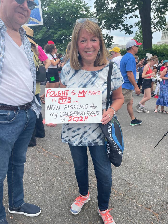 A woman in a tie-dye shirt holds up a sign reading &quot;Fought for my rights in 1973... now fighting for my daughter&#x27;s rights in 2022!!&quot;