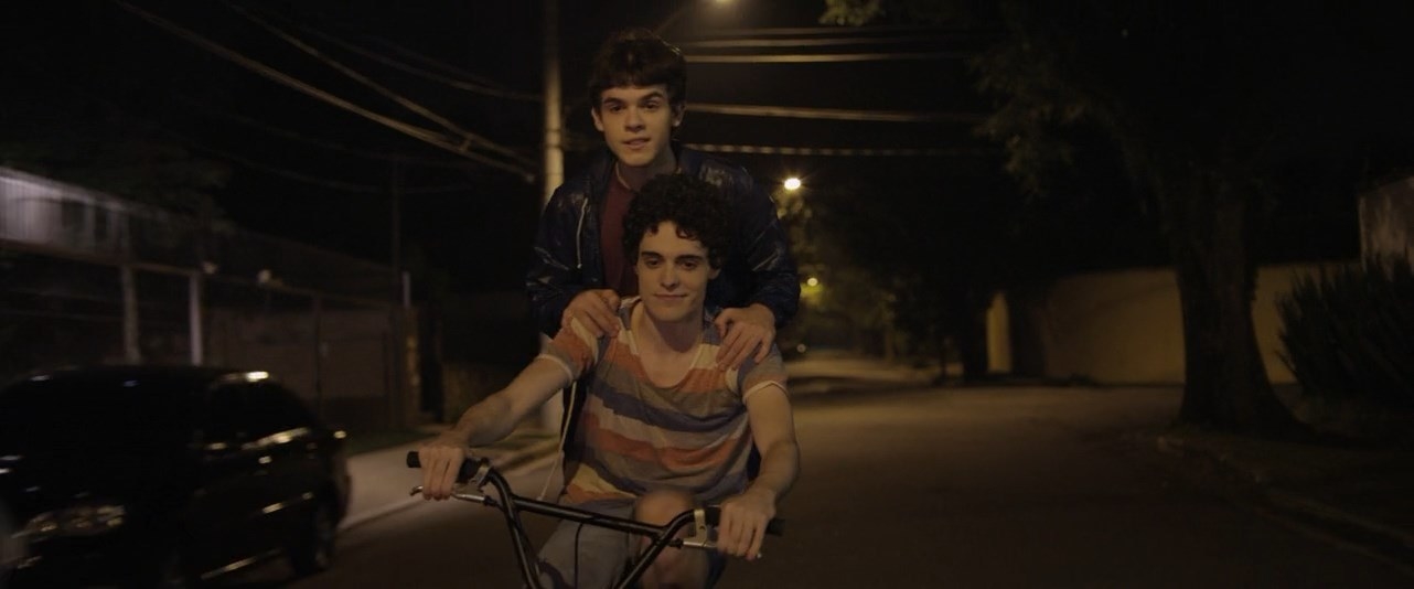 Gabriel (Fábio Audi) riding a bicycle with Leonardo (Ghilherme Lobo) behind him