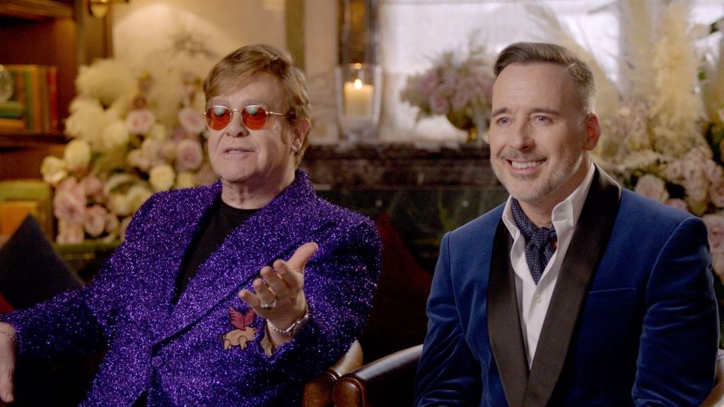 Elton John and David Furnish sitting next to each other in a room with flowers behind them
