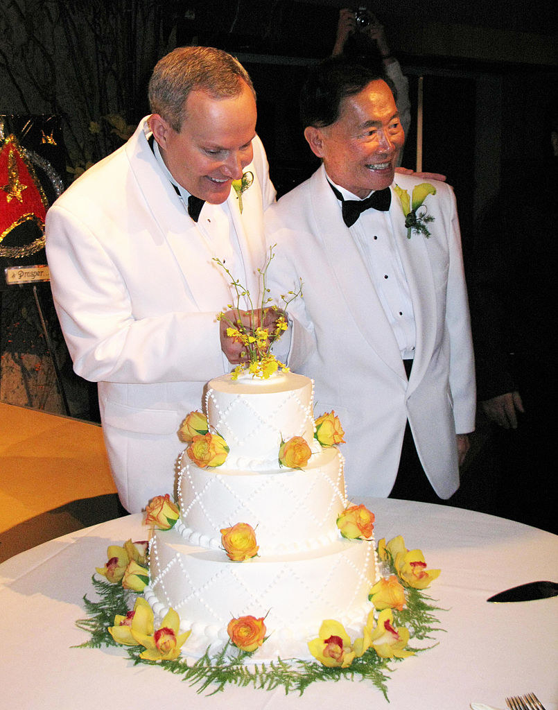 Brad and George Takei wearing white tuxes and standing by a wedding cake