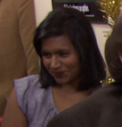 Mindy Kaling stands at an event, smiling softly, wearing a casual cap sleeve top