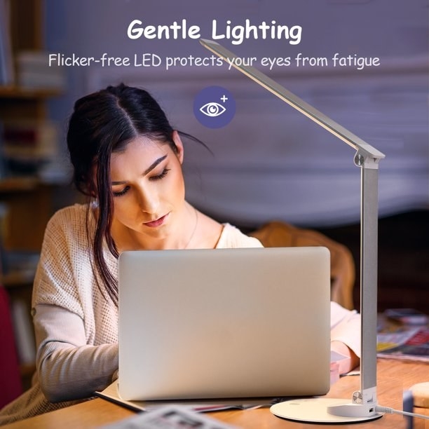 Model sitting at desk and working on computer with the lamp illuminating the space with gentle lighting. text reads &quot;flicker free LED protects your eyes from fatigue&quot;