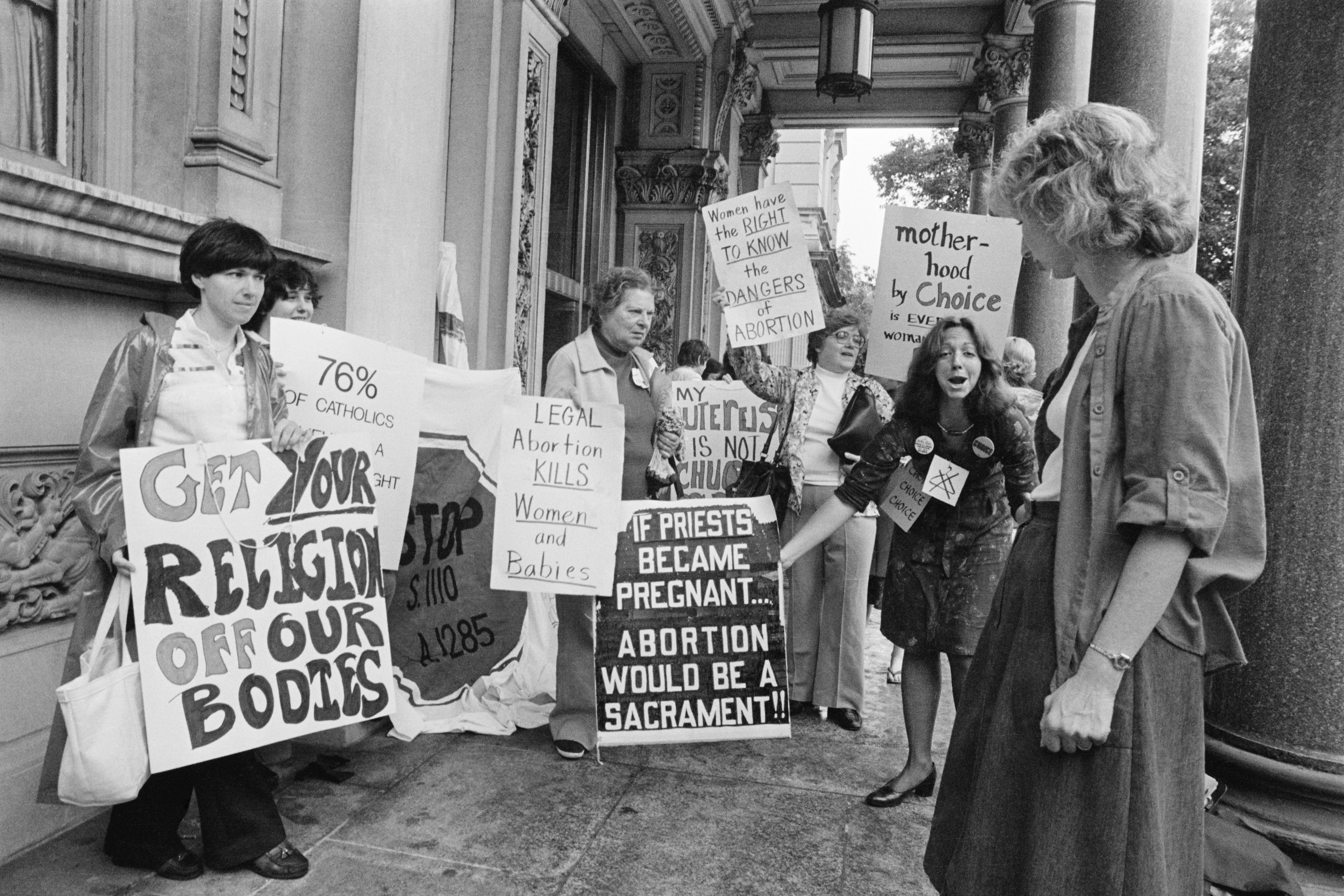 A woman stands near protesters carrying signs reading &quot;if priests became pregnant, abortion would be a sacrament&quot; and &quot;get your religion off our bodies&quot;