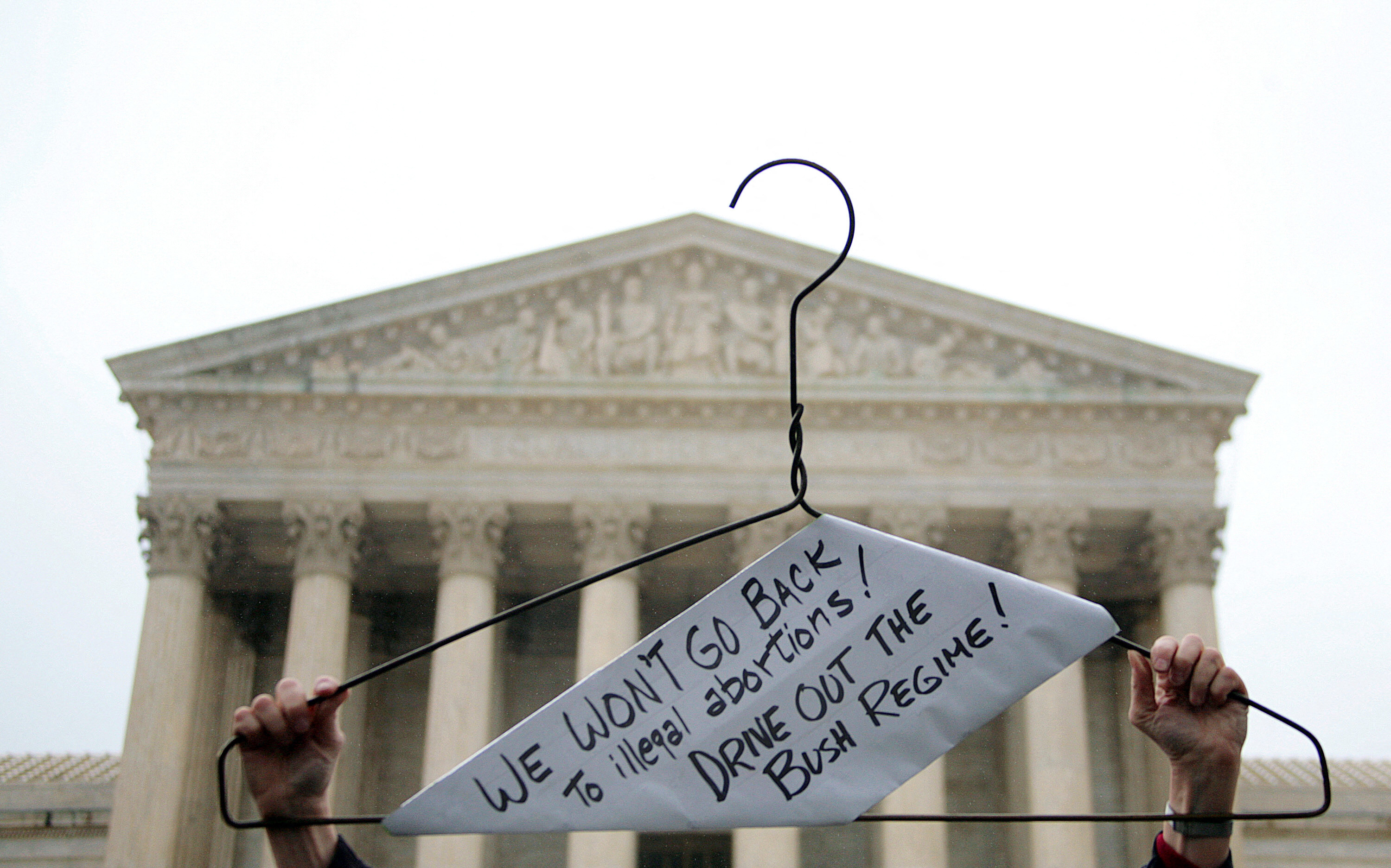 A coat hanger in front of the Supreme Court is held up and labeled with a piece of paper reading &quot;we won&#x27;t go back to illegal abortions, drive out the Bush regime&quot;