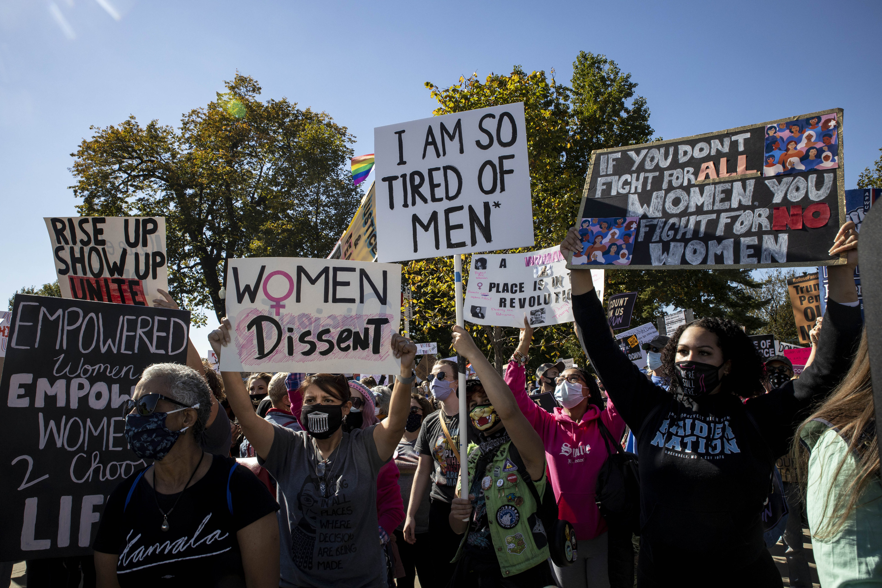 Protesters stand in a park and hold up signs reading &quot;I am so tired of men&quot; and &quot;women dissent&quot; and &quot;if you don&#x27;t fight for all women you fight for no women&quot;