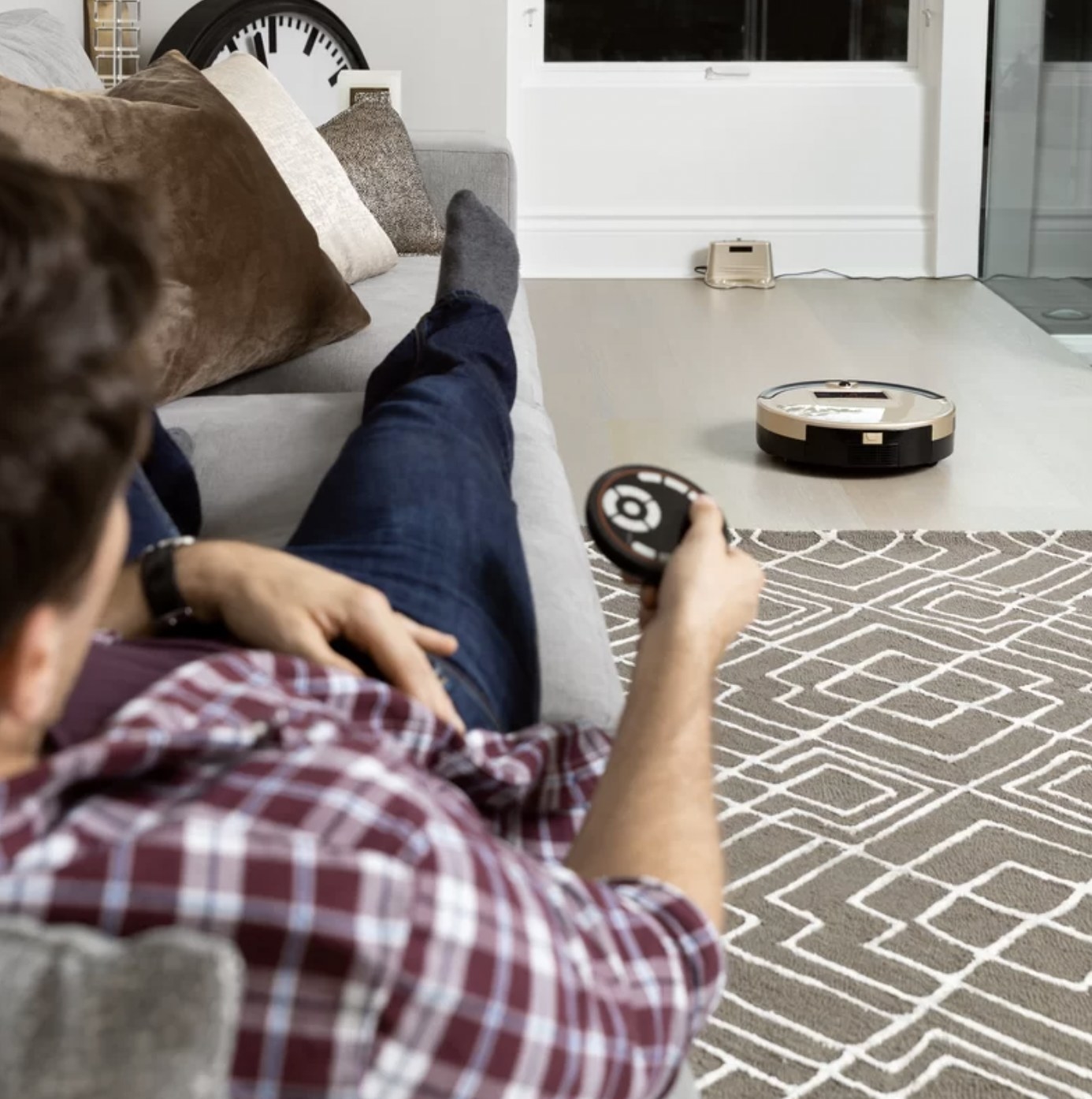 Model lounging on couch and using the remote to direct the robot vacuum on the floor