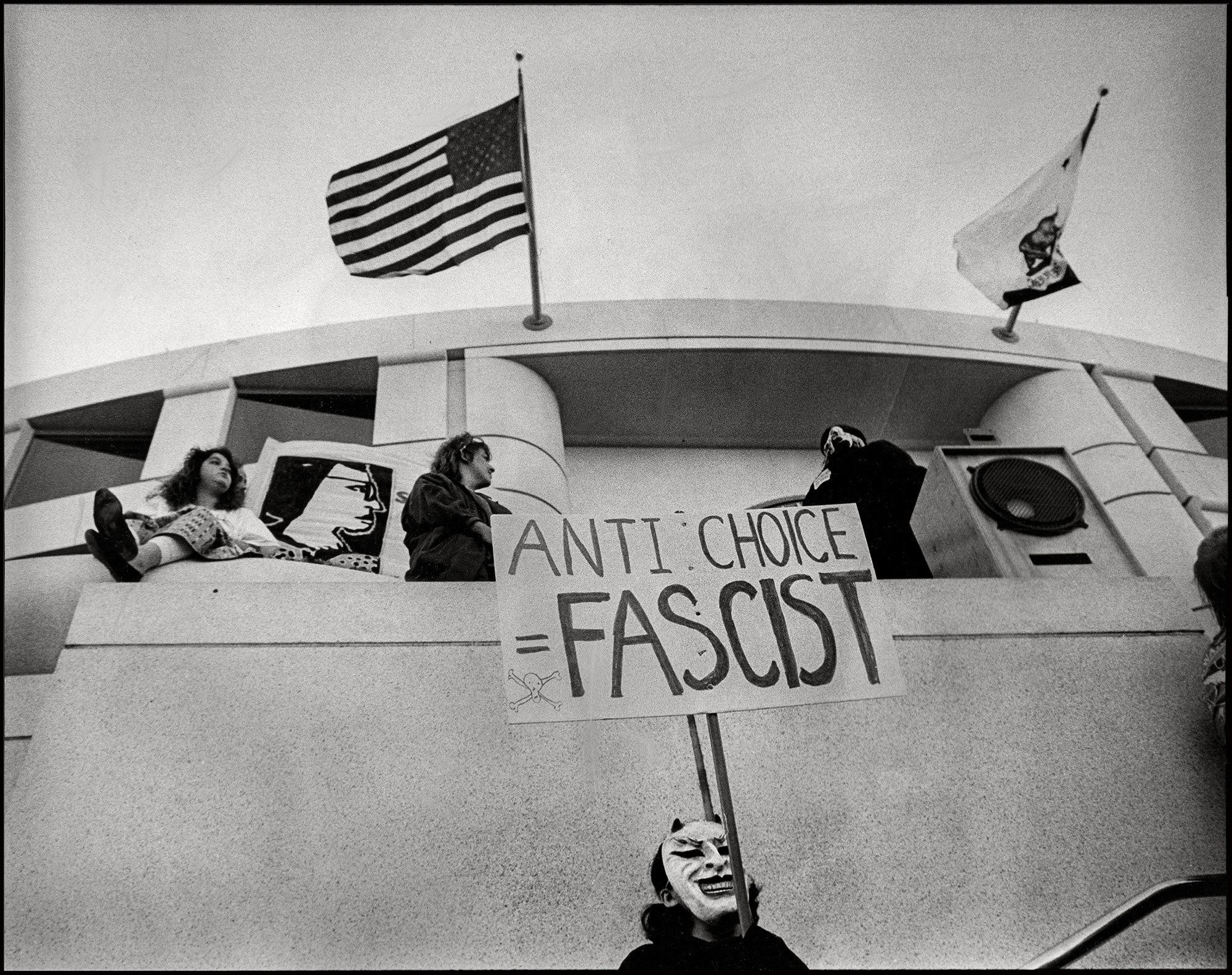 A demonstrator in a demon mask during a rally stands beneath California and US flags with a sign reading &quot;anti-choice equals fascist&quot;