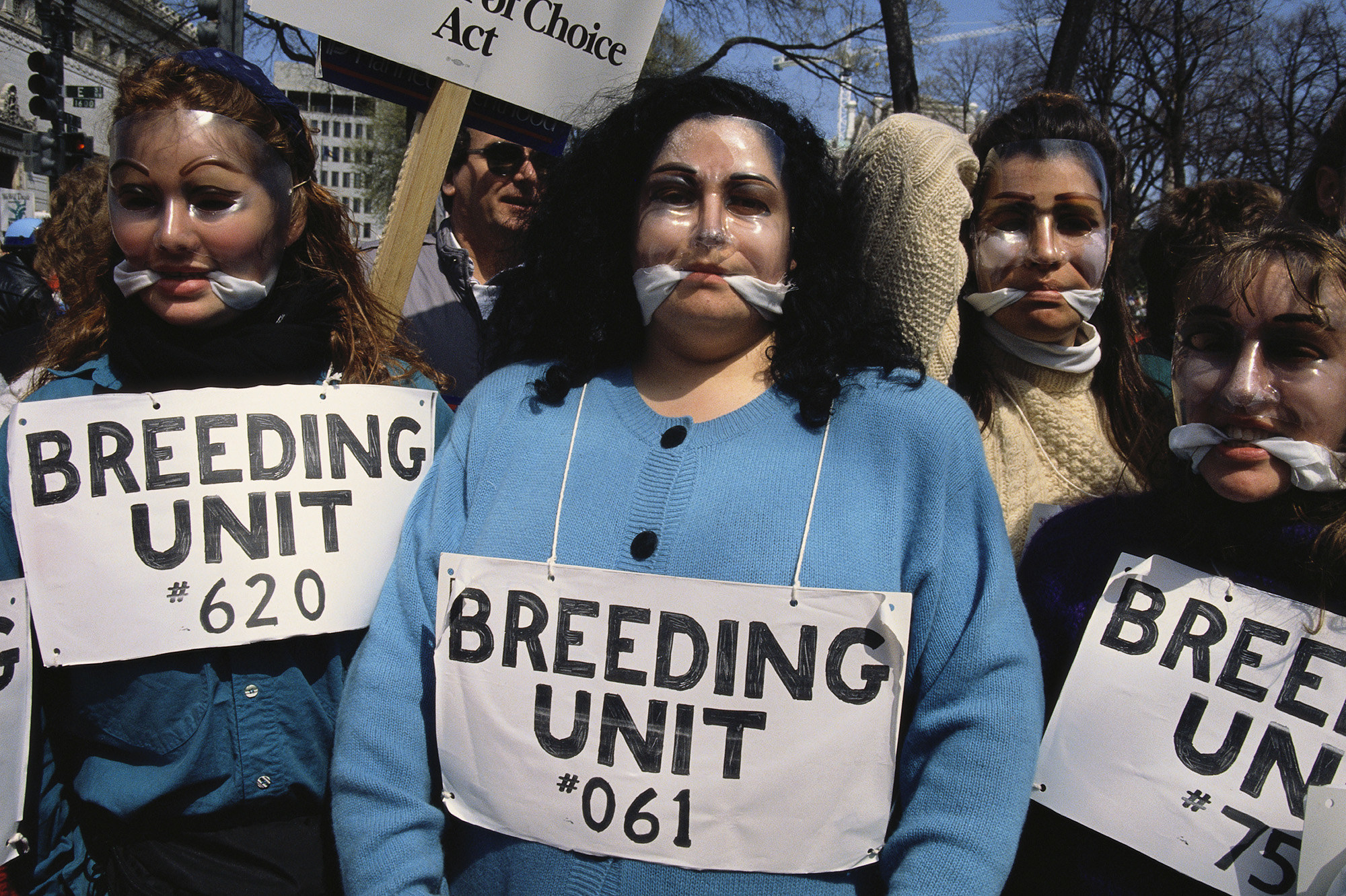 Women wear face masks and gags in their mouths with signs around their necks reading &quot;breeding unit&quot;
