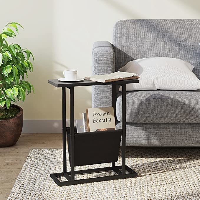 The end table with a cup of tea on top of it next to a couch in a living room with books in the sling