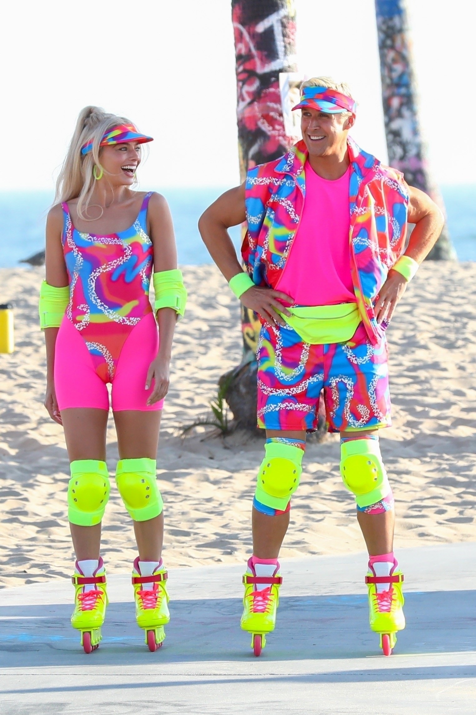 Margot still in rollerblades, now joined by Ryan; they&#x27;re both wearing matching neon outfits with bright knee pads and rollerblades